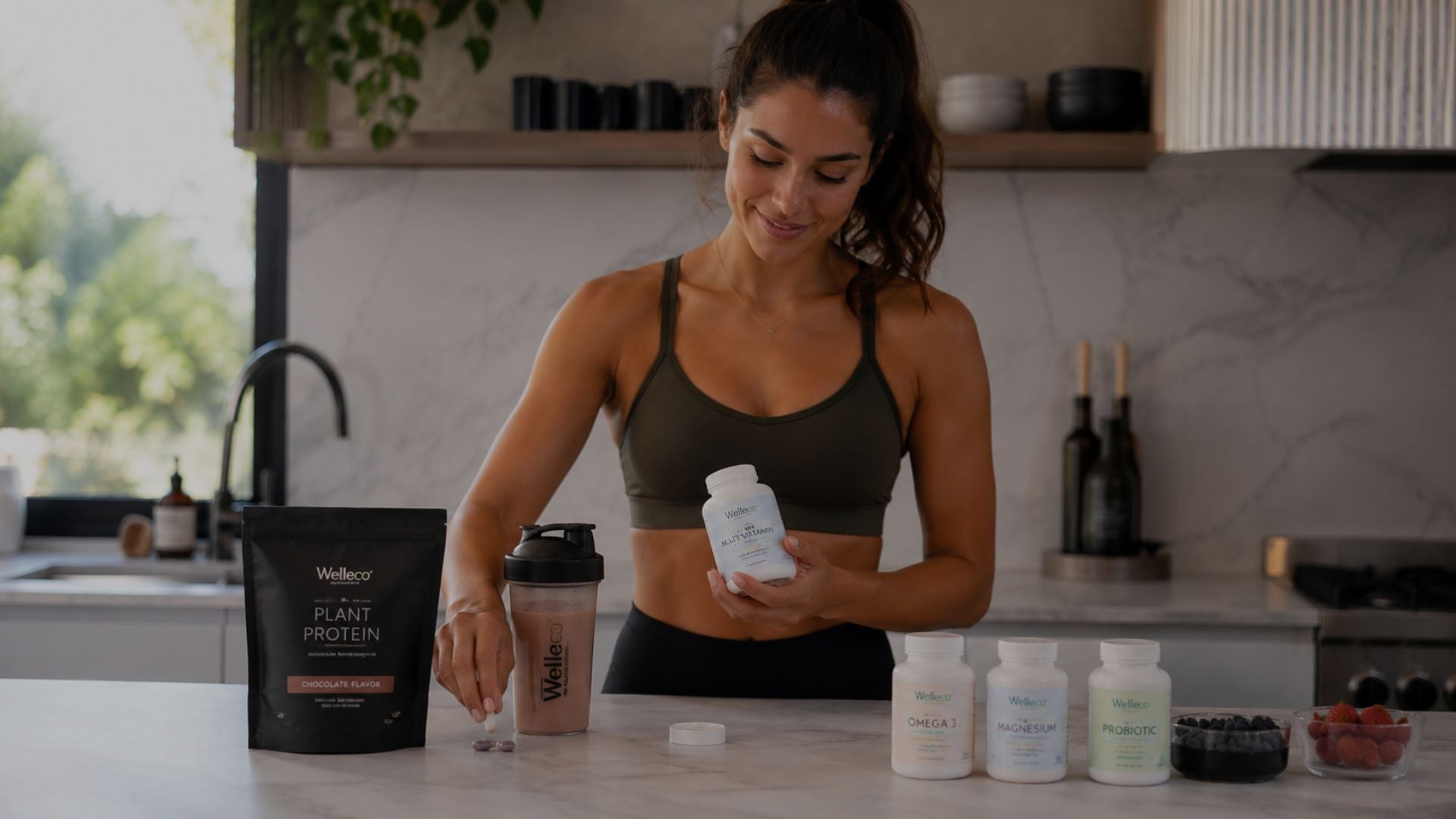 Smiling woman holding multivitamin bottle with protein shake and wellness supplements in kitchen