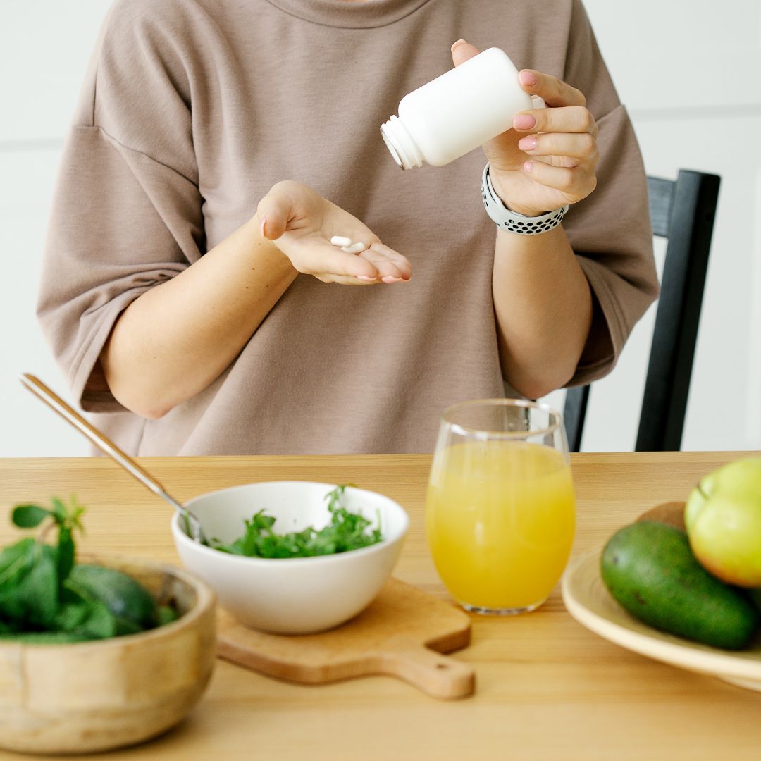 Woman pouring daily vitamins supplements into hand with healthy breakfast juice and greens on table